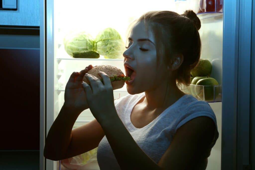 Femme mange un sandwich près du frigo ouvert.